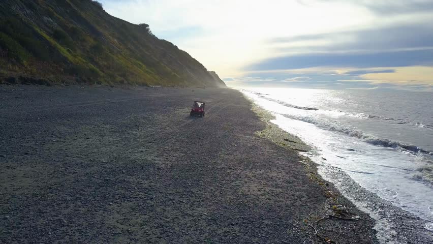 Cinematic aerial drone tracking shot of ATV or All Terrain Vehicle going on shore next to the crashing waves and the cliffs in Deep Creek State Park in Ninilchik, Alaska