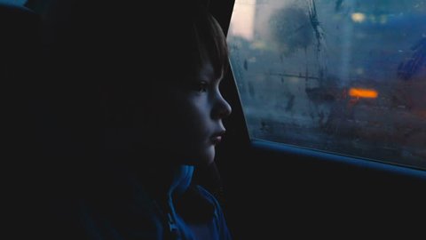 Atmospheric shot of little 4-6 year old thoughful Caucasian boy looking out of foggy car window in dusk dark evening.