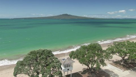 Aerial: Pohutukawa trees, lifeguard house and cyclist. There is a view across the Waitemata Harbour out to Rangitoto Island. Auckland, New Zealand. 15 November 2018