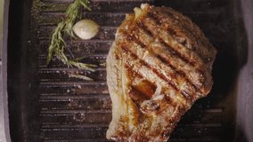 a beef steak is roasting with rosemary and garlic on a hot black pan close up view from above slow motion video in 4K