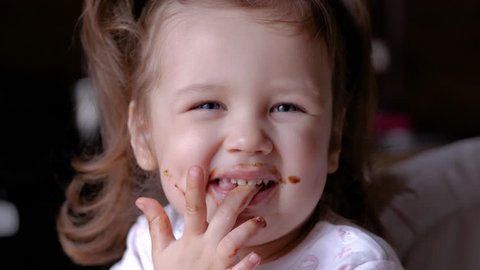 Closeup view of funny dirty small girl after eating chocolate. Brown lips and teeth. Child licking fingers.