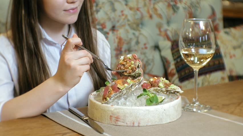 A beautiful blond woman eats a dish of raw meat tar tar in a seashell, uses a cutlery fork, drinks white wine in a restaurant. Hands close up