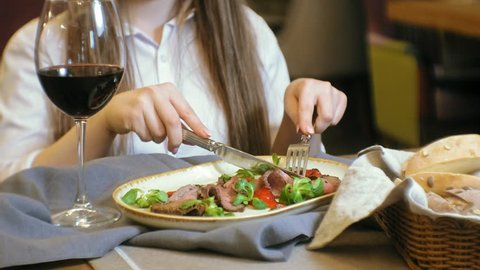 Beautiful blond woman eating meat dish, using cutlery, knife and fork, drink red wine in a restaurant. Beef meat dish, roast beef. Hands close up