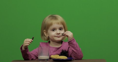 Girl sitting at the table and eating chocolate, cookies and drinks cacao. Happy three years old girl. Cute girl smiling. 3-4 year old blonde girl. Make faces. Green screen video. Chroma Key