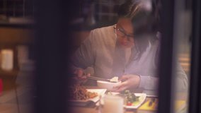 View Through window glass: Short-haired, glasses-wearing Chinese women are having dinner in restaurant, she looked down at cell phone and smiled, Businesswoman with mobile phone lifestyle 4k clip