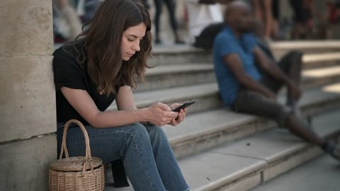 Timelapse of woman with smartphone sitting on the stairs in Paris texting. Caucasian young woman with dark hair wearing jeans and black shirt. Crowds passing by.