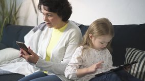 Woman is sitting on sofa and touching phone screen while child using laptop for playing or computing at home. Digital wireless electronic devices for network and game. Mother and daughter is working
