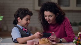 Close up curly mother with cute mixed race boy watching funny video on smart phone after festive breakfast in home kitchen. Laughing family celebrating together Mother's day