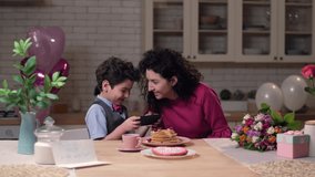 Happy mother and cute joyful preadolescent son watching amusing video clips on cellphone during holiday breakfast in festively decorated kitchen. Laughing family having fun together at Mother's day