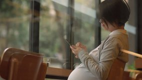 Side view of one pregnant Asian woman using a mobile phone in the cafe by the window. pregnant female lifestyle with mobile technology 4k clip