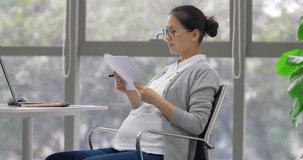 Side view of one Chinese pregnant businesswoman wearing glasses is working at her laptop on the table in the office, making notes on the documents, Asian working woman in pregnancy  4k clip