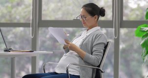 Chinese pregnant businesswoman working in front of laptop on the table in the office, making notes on the documents and touching her belly gently and softly. Asian working female in pregnancy 4k clip