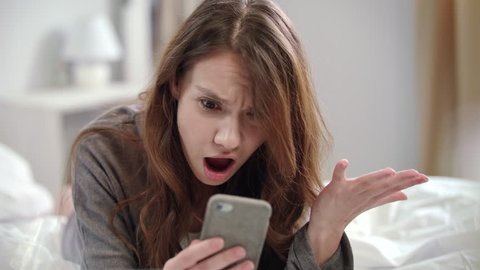 Shocked woman watching video news online on mobile phone at morning. Surprised woman face looking at smartphone in bedroom. Close up of young woman looking shocked video online on mobile phone