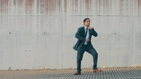 Cheerful and Happy Businessman is Actively Dancing on a Street Next to an Urban Concrete Wall. He's Wearing a Grey Suit. Sunny Day.