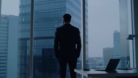 Back View of the Thoughtful Businessman wearing a Suit Standing in His Office, Contemplating Next Big Business Deal, Looking out of the Window. Business District Panoramic Window View