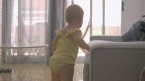 Back view of one year old baby girl walking at home leaning on the sofa. Home video imitation