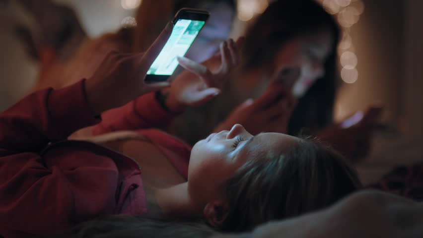 Happy teenage girls lying on bed using smartphone texting on social media browsing online having fun sleepover on weekend night 