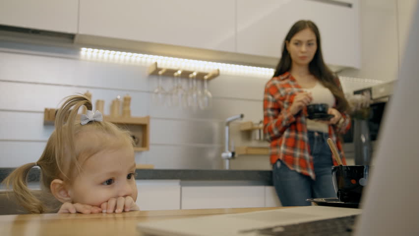 Female toddler watching cartoons on latpop in the kitchen and her mother looking to her. Little girl standing at the wooden table with computer on and woman with long hair drinking tea and smiling on