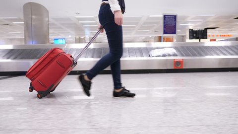 Woman hold trolley case handle and quickly walk to exit across luggage reclaim hall. Lower half tracking camera motion, side view of legs and wheeled suitcase, baggage carousel slide on background