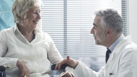 Professional doctor and nurse talking to a smiling senior female patient on a wheelchair and comforting her, the geriatrician is holding her hands - Powered by Shutterstock - Get 15% off with code: PIKWIZARD15