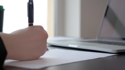 Female hand writing paper document with pen on working table on laptop background. Close up business woman writing document on desktop in office. Fill in form blank sign finance document. Journalist