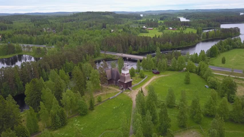 Aerial, drone shot, towards the famous, wooden church, in the middle of a lake, a road and green trees, on a cloudy, summer day, in Petajavesi, Middle Finland