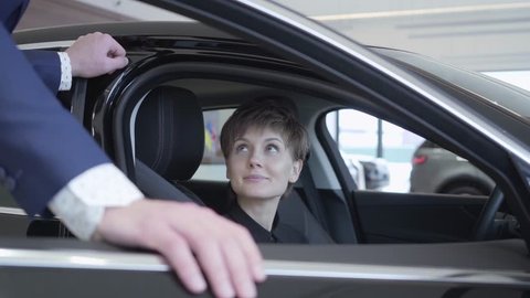 Cute young woman with short hair sits in modern car talking with unrecognizable salesman. Successful woman examines new auto. Professioal salesman in suit consulting customer. Concept of buying