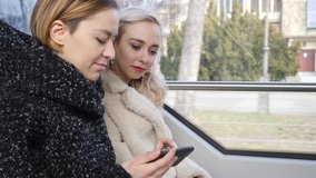 Two pretty young women watch smart phone social media video sitting in tram city transport