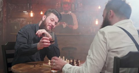Two young hipsters clinking glasses of whisky and drink at a chess game in a pub. A conversation between two friends in a bar.
