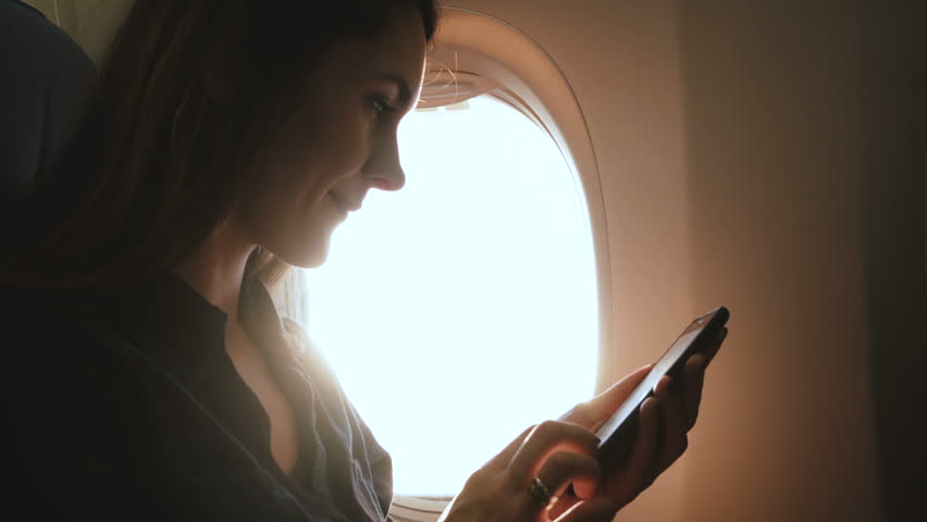 Amazing close-up shot of young relaxed woman using smartphone app and drinking tea on airplane flight sunny window seat. Royalty-Free Stock Footage #1024974104