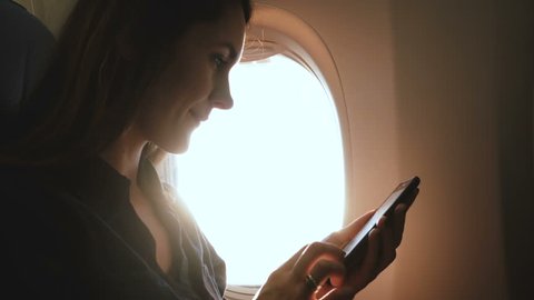 Amazing close-up shot of young relaxed woman using smartphone app and drinking tea on airplane flight sunny window seat.
