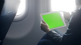 Young brunette hair woman wearing in suit or formal wear holding portative tablet with copy space display in hands. She sitting near window in airplane and using app to discussing about journey