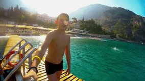 Teenage boy dives into the blue sea from the bridge and makes selfie video.   Happy kid is diving from the wooden bridge into water. Slow motion. Mediterranean sea. Beautiful landscape. Turkey.