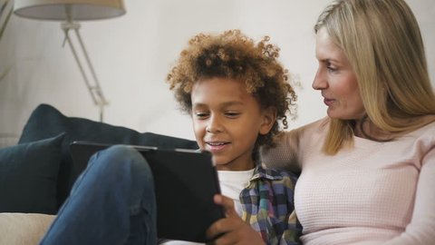 Glad and positive woman mom sitting on comfort couch inside bright light living room with her glad and excited son. They looking at tablet together, making wide beaming smile and clapping in palm