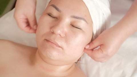 Close-up. Face massage of a middle-aged Asian woman in a beauty salon. The movement of the fingers of the beautician's hands from the chin to the eyes. Raw video without color correction.