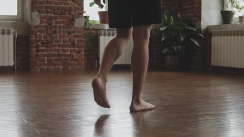Young man in shorts enter yoga studio with wooden floor and big windows in slow motion. Close-up of male with yoga mat
