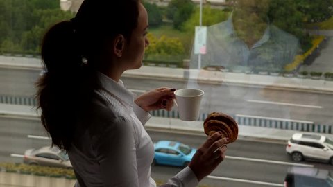 Woman stay fixed against window, hold cup of hot coffee and croissant, slow motion shot. Business lady take break to drink and eat pastry, stay lost in thought, stare outdoors. Car traffic seen down