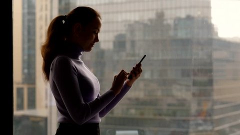 Young adult woman stay with phone in hands against room window, type message. Blurred background, office building seen outdoors. Dim light, shaded silhouette of lady
