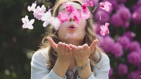 Woman Enjoying Spring Blossom. SLOW MOTION. Happy Smiling Girl blowing on rhododendron flower petals, making them fly to camera. Sunny Spring Outdoors Activity, Lens Flare.