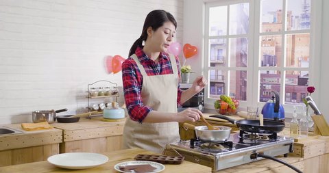 Happy asian young woman singing dancing while cooking in modern wooden kitchen. female in apron with love preparing valentine day surprise. girl with wooden spoon stirring dark chocolate smelling.