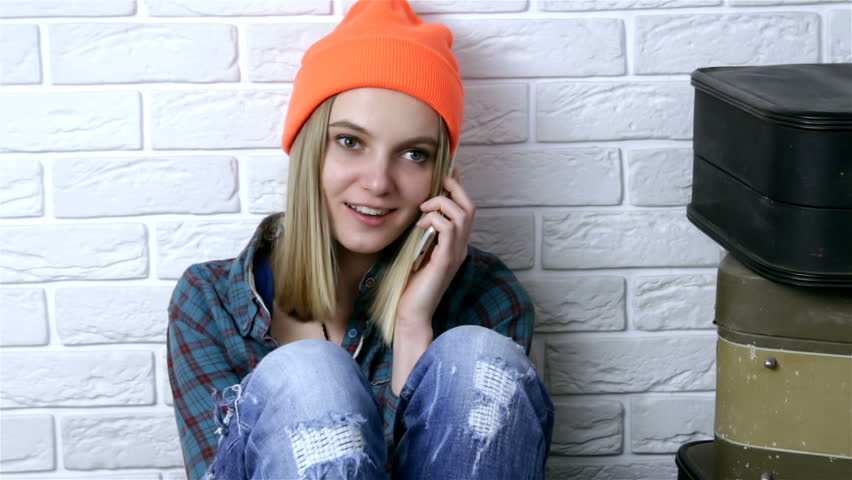 Fashion hipster  stylish young woman in orange hat using smartphone over white brick wall