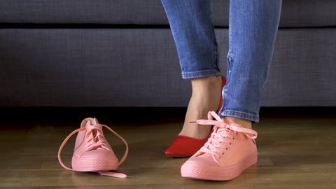 Slim woman in jeans on gray couch and takes off sexy red heels and puts on comfortable coral sneakers