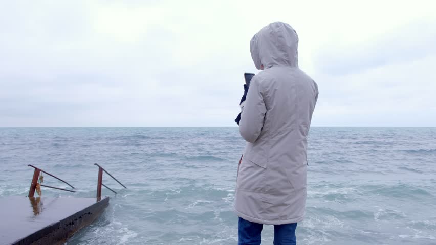 Woman in a white jacket takes a mobile phone storm waves standing on embankment. Back view.