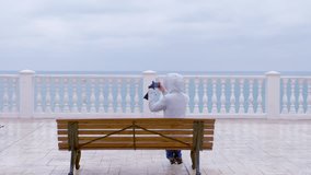 Woman in white jacket and hood shoots sea waves video on smartphone sitting on the bench on beautiful terrace with sea view on waterfront. Back view.