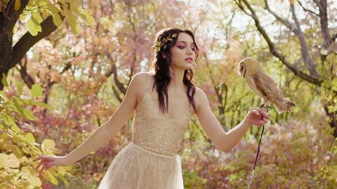 spring nymph of nature and forest fairy, dark-haired beauty in long dress holding cute little owl on her arm, fairy-tale princess with delicate pink make-up, model posing in magical forest
