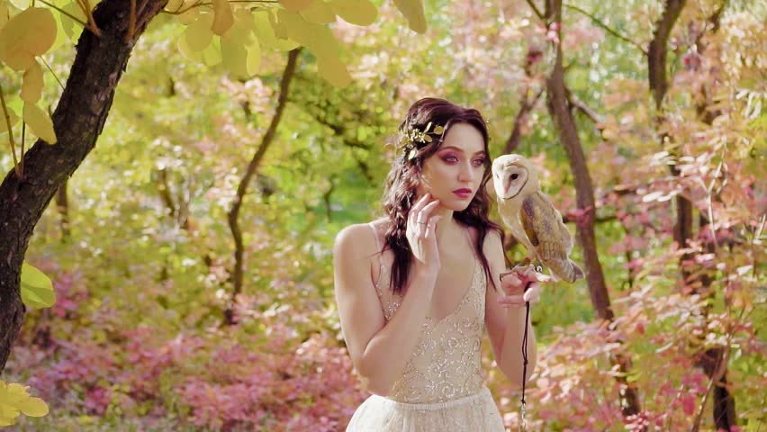 the spirit of the spring forest holds a gorgeous little cute bird on her hand, a fantasy nymph with a golden wreath on dark curly hair girl model in a long beige dress posing with a tender owl