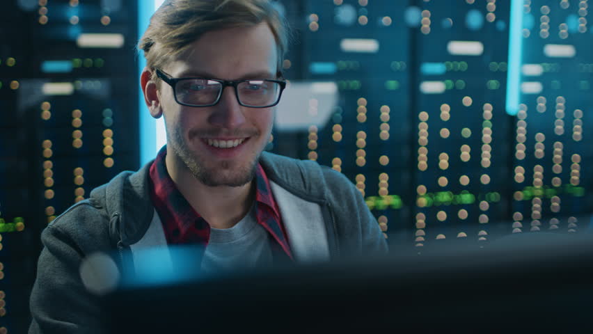 Portrait of a Smart Focused Young Man Wearing Glasses Working on a Desktop Computer. In the Background Technical Department Office with Functional Data Server Racks. | Shutterstock HD Video #1025052656
