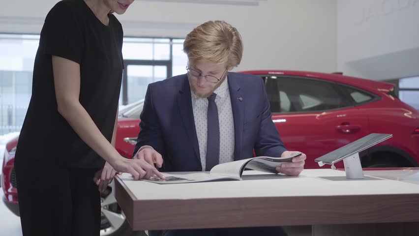 Unrecognizable woman stands near confident seller man sitting at the table. Salesman discuss choosing of car with customer turning book page in motor show