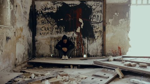A ,troubled young man wearing a hood in an empty, wrecked,abandoned building, in mental suffering agony and remorse.Slider, tracking motion.