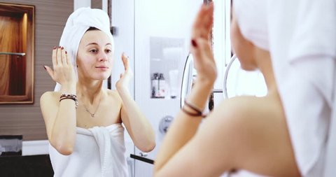 Young Woman Applying Face Cream. SLOW MOTION. Woman performing Beauty rituals after shower in front of bathroom mirror. Healthy Skin hydration and moisturising.
